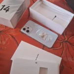 Unboxing do OPPO Reno 14F: caixa aberta revelando o celular azul Iridescent Mermaid, carregador SuperVOOC e acessórios sobre mesa vermelha.