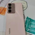 realme c75x vale a pena em 2026: foto real da traseira do smartphone na cor rosa conectado ao cabo do carregador sobre desenhos.