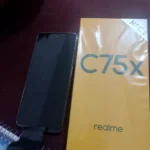 realme c75x vale a pena em 2026: aparelho com tela desligada posicionado ao lado da caixa oficial amarela da marca com selo NFC.