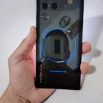 "Visão traseira do smartphone gamer RedMagic 11 Pro com design transparente, luzes RGB azuis e indicação da zona de carregamento sem fio de 80W, segurado por uma mão