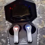 Fone bluetooth Altomex A607 encaixado na case de carregamento com LEDs acesos em vermelho e azul sobre uma bancada de pedra escura.
