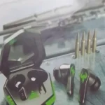 Fone gamer bluetooth Altomex A607 preto com detalhes em LED verde fora da case de carregamento aberta sobre uma superfície temática com desenho de munições.