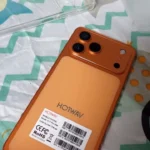 Traseira do smartphone HOTWAV A17 Pro Max na cor Laranja Sunset (Sunset Orange). Mostra o módulo de câmera tripla no canto superior esquerdo e uma etiqueta branca na parte inferior com códigos IMEI e o modelo "A17 Pro Max". Uma capa transparente está deitada no fundo em um tecido estampado.