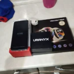 Vista superior do kit de unboxing do HOTWAV A17 Pro Max. O smartphone está em pé sobre sua base vermelha com a tela "About Phone" ligada. Ao lado, a caixa preta do smartwatch URANYX e a embalagem maior com texto "VEM COM 7 TIRAS" e imagem do relógio. Um cabo de carregamento e uma garrafa rosa estão ao fundo.
