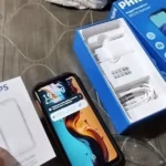 Unboxing do smartphone robusto Philips S8320 Xenium. A imagem mostra o aparelho ligado com wallpaper colorido, a caixa azul aberta contendo o carregador de parede e cabo USB brancos, e o manual de guia rápido ao lado.