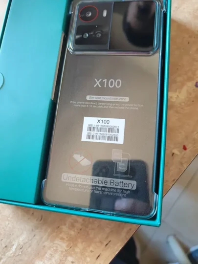 Traseira do smartphone Cubot X100 na cor cinza escuro, ainda com película de proteção, dentro de sua caixa de varejo verde azulado aberta. Detalhes como o módulo de câmera e adesivos de IMEI são visíveis.