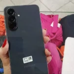 Mão segurando a parte traseira do Galaxy A56 na cor grafite, destacando as três câmeras verticais e o acabamento premium.