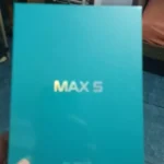 Fotografia amadora da caixa de embalagem de um smartphone Cubot Max 5, de cor turquesa com texto dourado, segurada pela mão de um comprador.