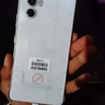 Fotografia amadora da parte traseira de um smartphone Cubot Max 5 branco com padrão geométrico, em uma capa transparente, segurado pela mão de um comprador e conectado a um cabo de carregamento USB-C.
