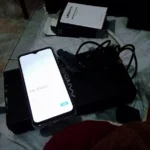 Smartphone Oukitel C3 ligado exibindo a tela de boas-vindas sobre a caixa do produto e acessórios.