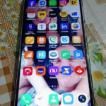 Foto em close de um smartphone com a tela ligada, exibindo um papel de parede de um casal sorrindo. A grade de aplicativos mostra ícones de bancos brasileiros como Itaú, Bradesco, Nubank, Caixa, Banco PAN e outros, além de redes sociais como Facebook e WhatsApp. O aparelho está sobre uma toalha de mesa floral.