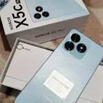 Uma visão de unboxing do smartphone Honor X5c Plus na cor azul claro metálico. O aparelho está em primeiro plano, com a parte traseira voltada para cima, mostrando seu módulo de câmera tripla circular e o flash LED. Abaixo dele, a caixa branca original do produto com o texto 'HONOR X5c Plus', 'NFC' e '5260mAh' está visível e aberta.