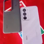 Descubra se o Redmi 15 é bom para jogos nesta foto da traseira do modelo Sandy Purple de 256GB com sua câmera de 50MP, posicionado ao lado da capinha de silicone oficial sobre a mesa vermelha de testes.