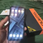 Foto real da mão do cliente shopee segurando o celular Oukitel G1 ligado ao lado da caixa oficial que destaca os 24GB de RAM e 256GB de memória.
