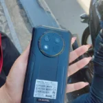 Visão traseira de foto real tirada por cliente do marketplace shopee do celular indestrutível Oukitel G1 em preto com detalhes em laranja no módulo de câmera Sony.