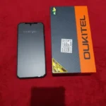 Smartphone Oukitel WP60 preto de frente com tela desligada ao lado de sua caixa de varejo laranja e preta sobre um fundo vermelho texturizado. Imagem real de unboxing para o review se o Oukitel WP60 é bom.